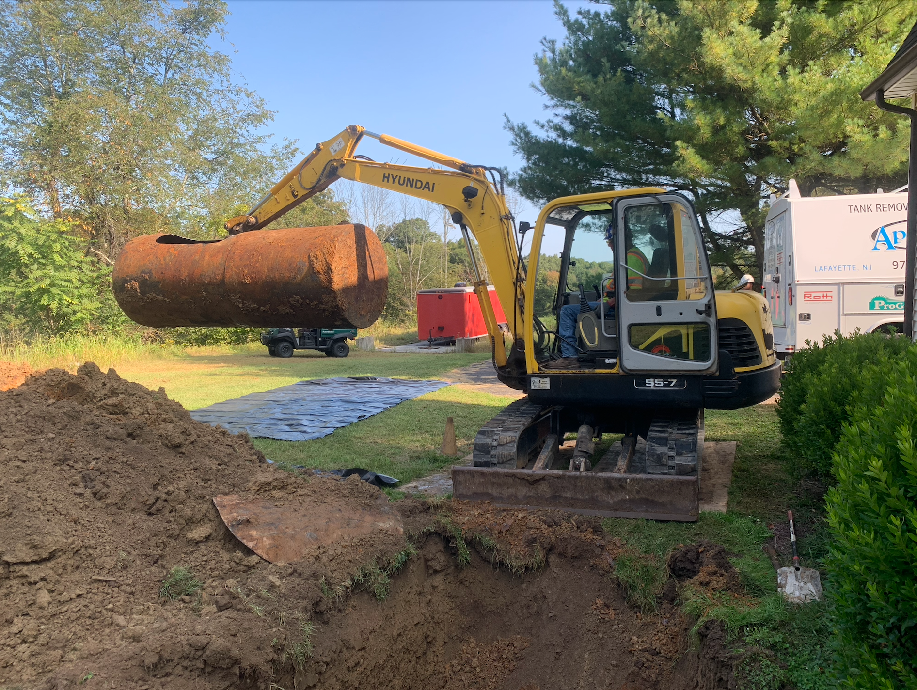 Excavator lifting a rusty underground storage tank from a yard with a dirt pile.