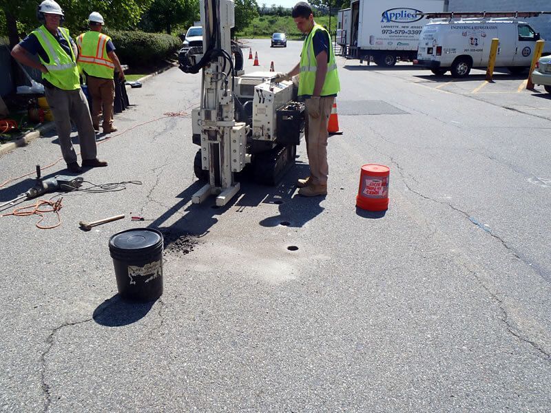Workers drilling pavement in a parking lot, using a machine; two observers nearby.