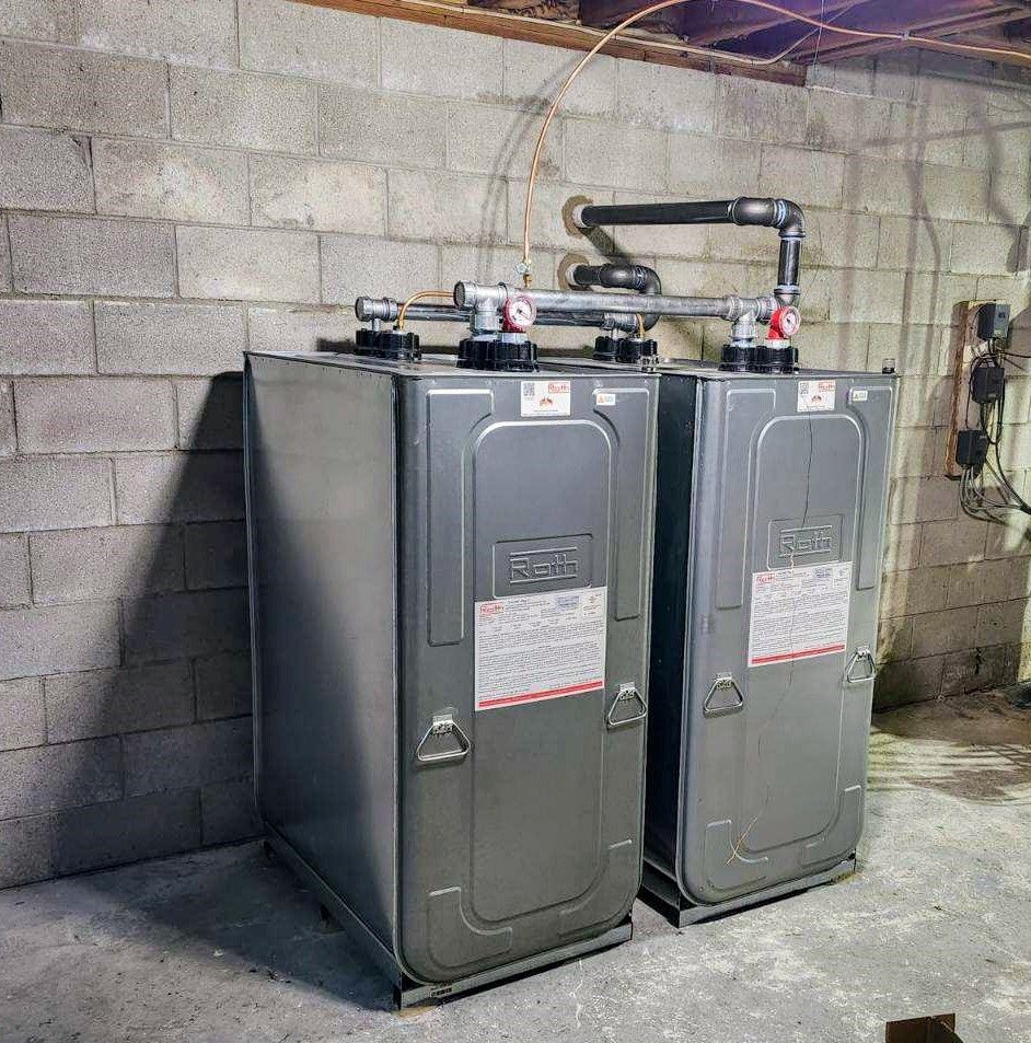 Two gray, rectangular furnaces against a gray concrete block wall in a basement.