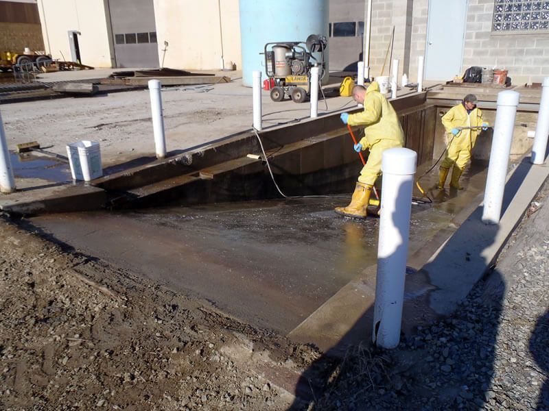 Two workers in hazmat suits spray a decontamination pad near a building with white pillars.