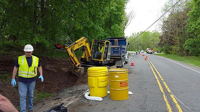 A man in a yellow vest is standing next to a yellow barrel on the side of a road.