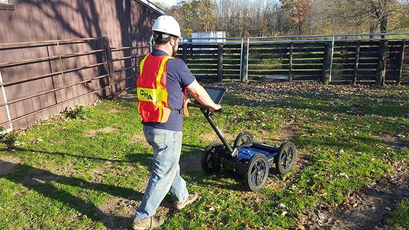 Man in safety vest using ground penetrating radar in a grassy area near a fence.