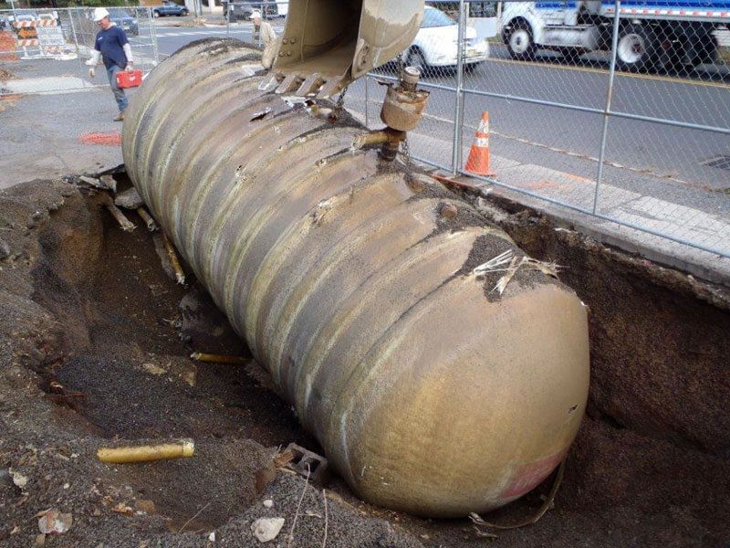 Excavator lifting a rusted underground storage tank from a dirt pit on a city street.