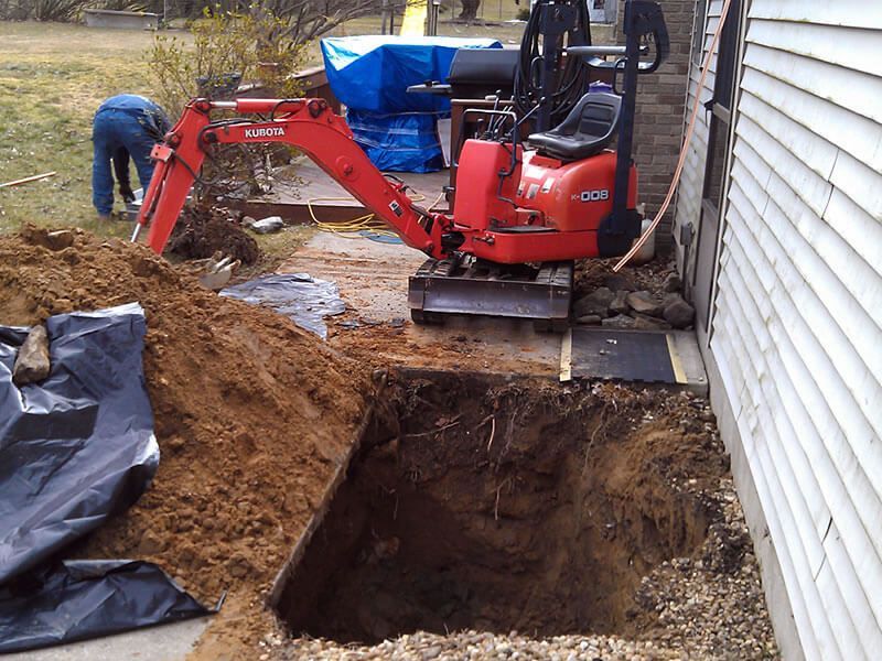 Red excavator digging a deep square hole next to a white building. A person works nearby.