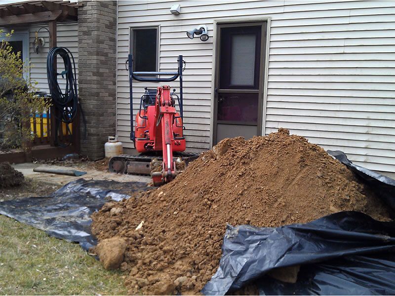 Red excavator digging near a house; dirt pile in foreground.