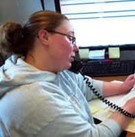 Woman on phone at desk wearing glasses and a light-colored hoodie, writing on paper.