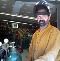 Welder wearing helmet and protective jacket smiles in a workshop with gas tanks.