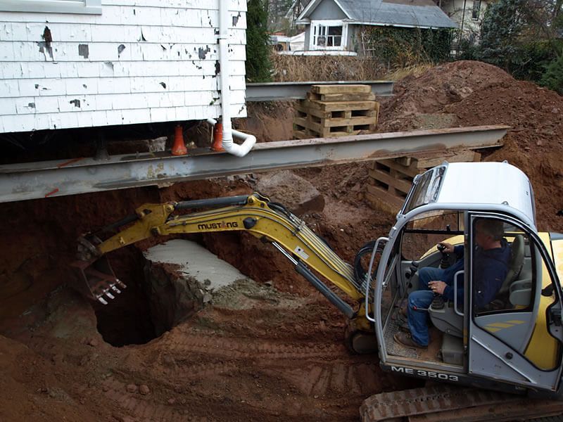 A man is driving an excavator in the dirt near a house