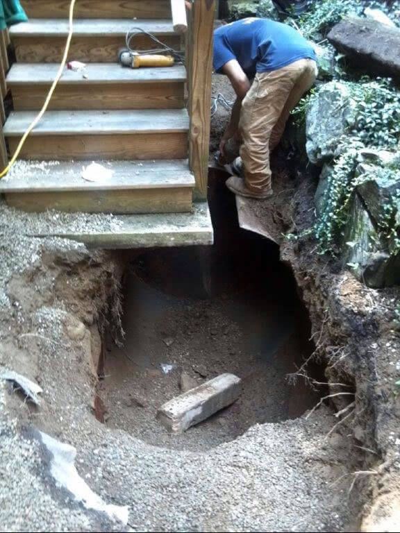 A person digs a hole near wooden stairs. Gravel, dirt, and rocks are visible in the setting.