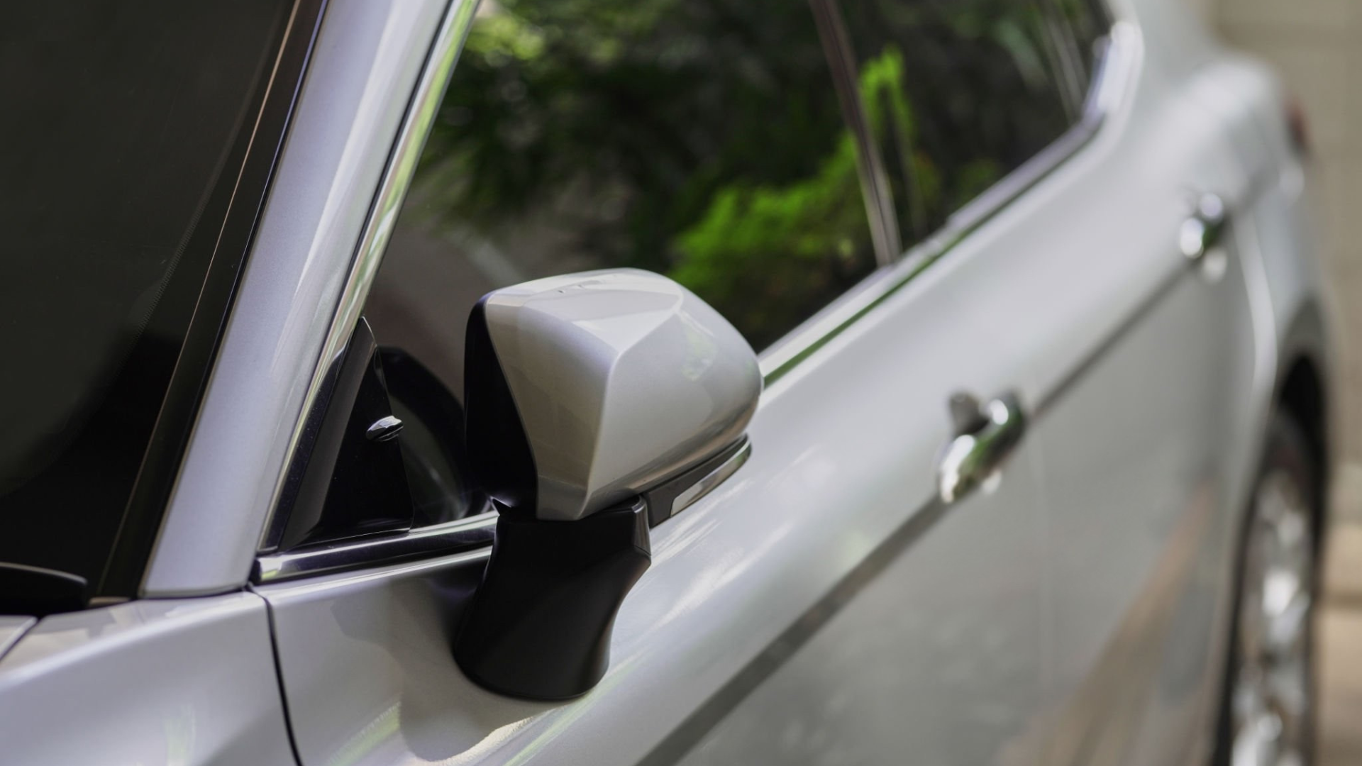 Silver car's side mirror and window. The mirror is rectangular, the window is tinted, and the door handle is visible.
