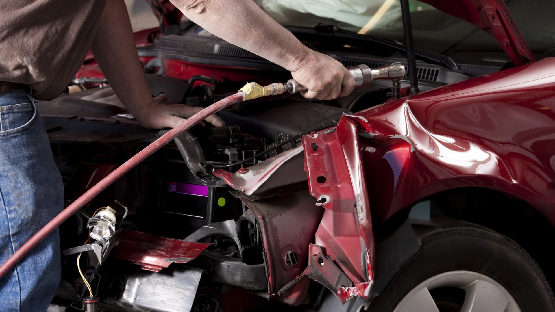 Mechanic repairing a heavily damaged red car in a garage. They hold a tool, working on the car's crumpled front.