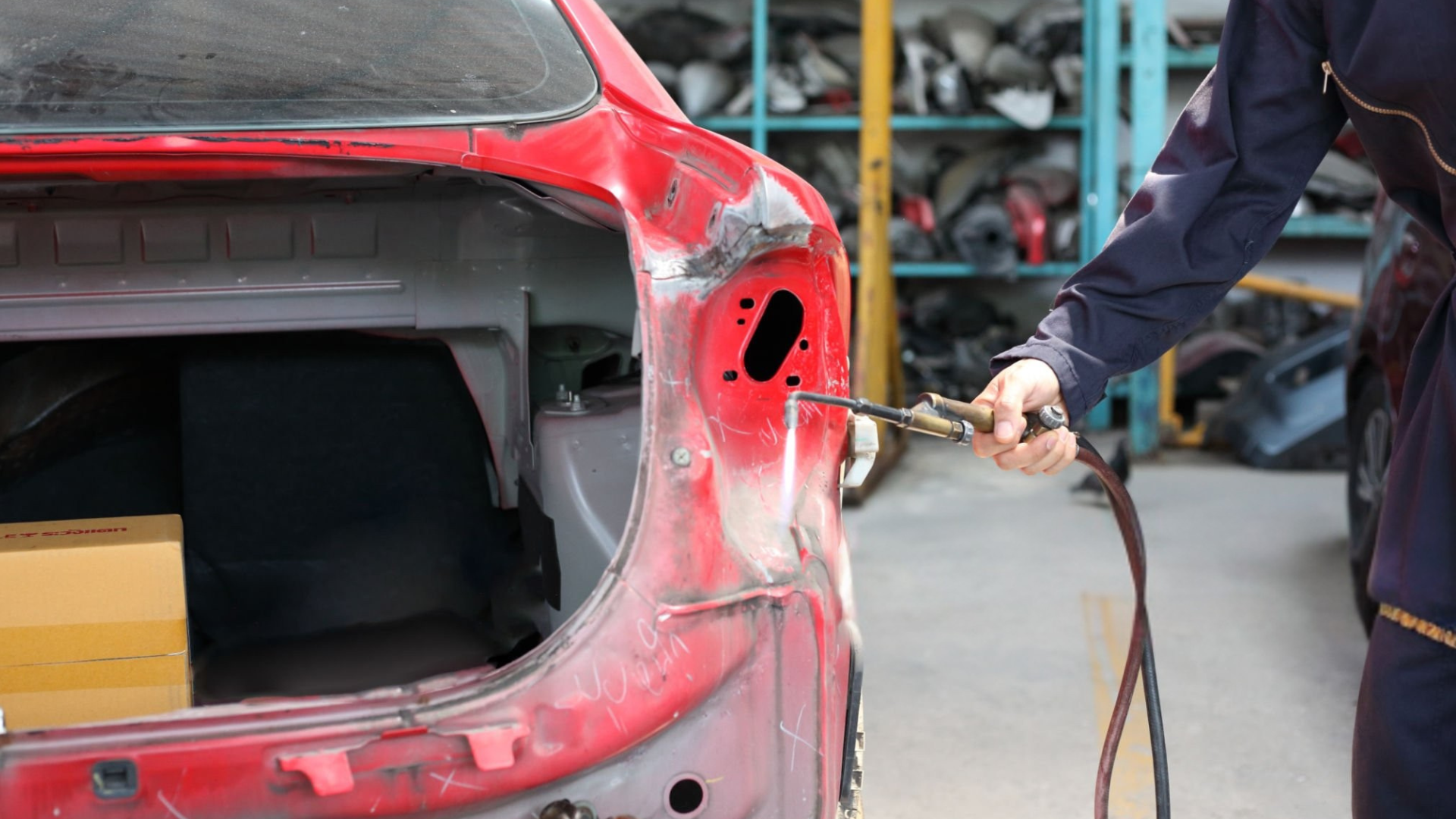 A person welds a car frame in a workshop; the car is red, and the person wears a blue jumpsuit.
