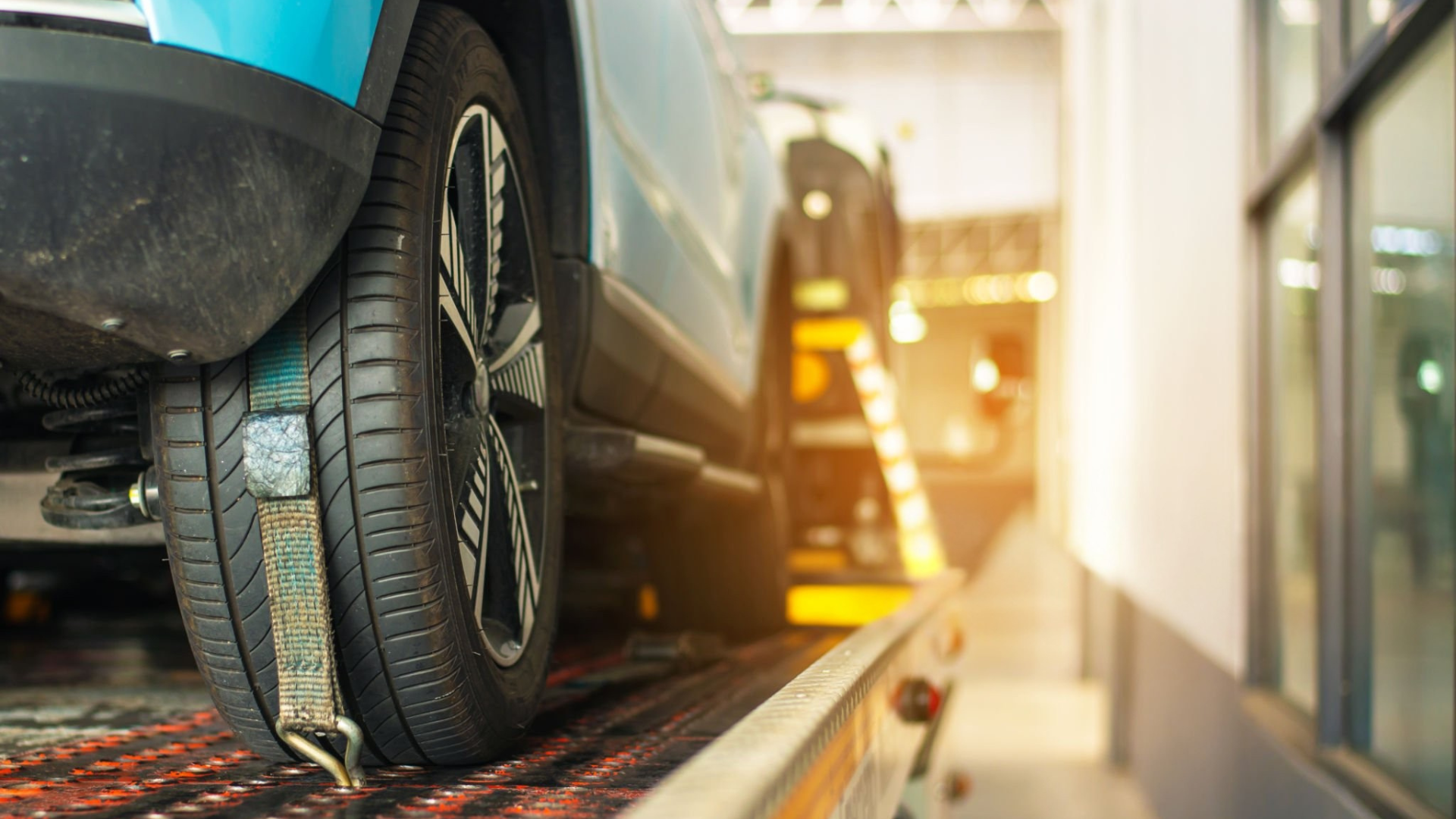 A blue car being towed onto a flatbed tow truck. The tire is secured with a strap, inside a repair shop.