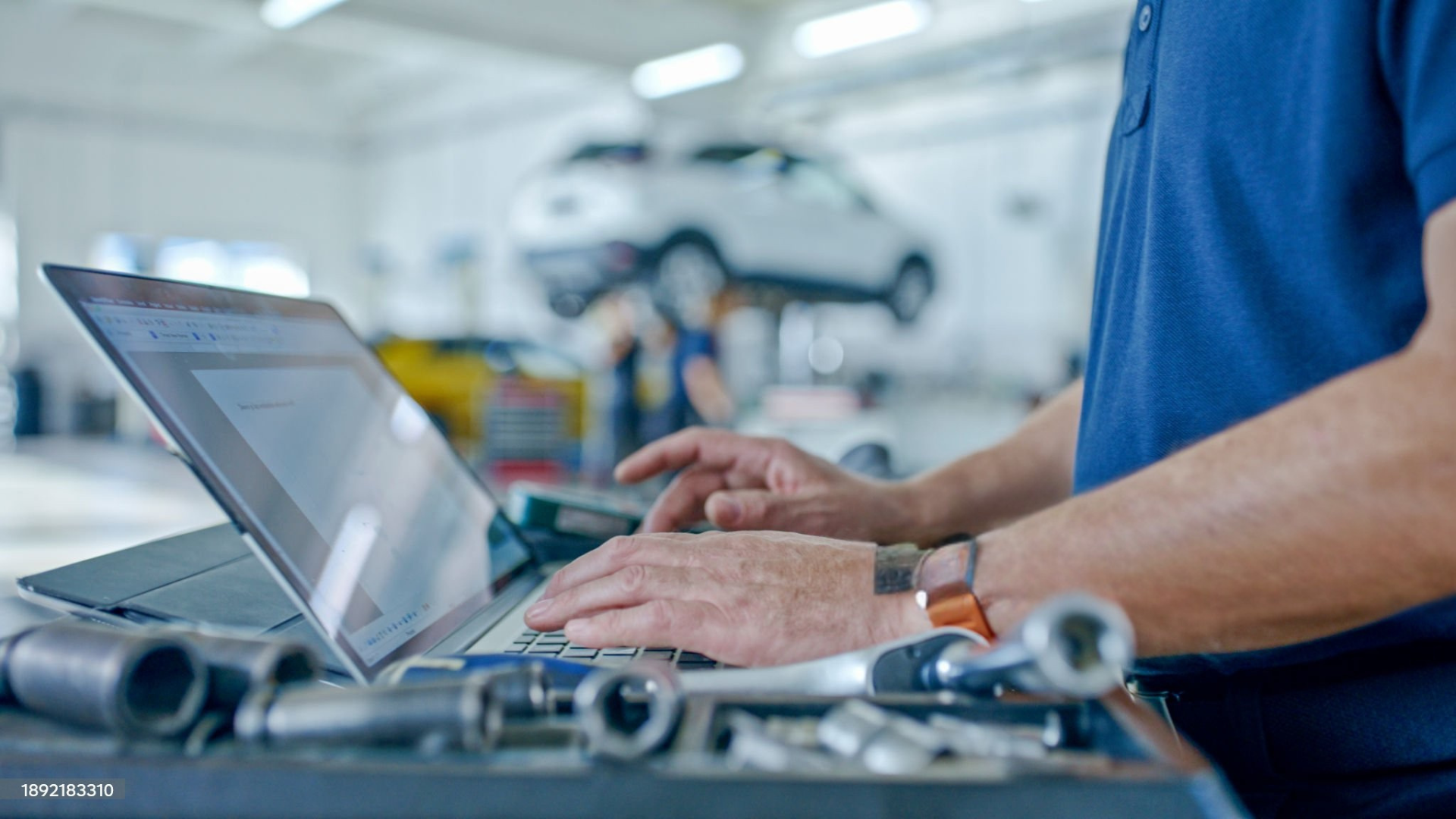 Mechanic using a laptop in a car repair shop, with tools in front and a car on a lift in the background.