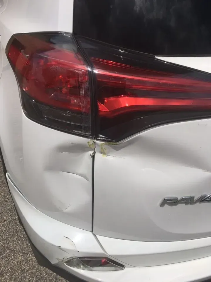 A white car with a damaged tail light is parked on the side of the road.