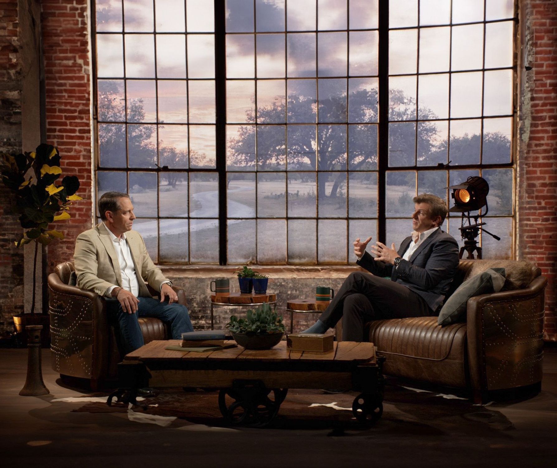 Two men are sitting in chairs and talking in front of a large window.