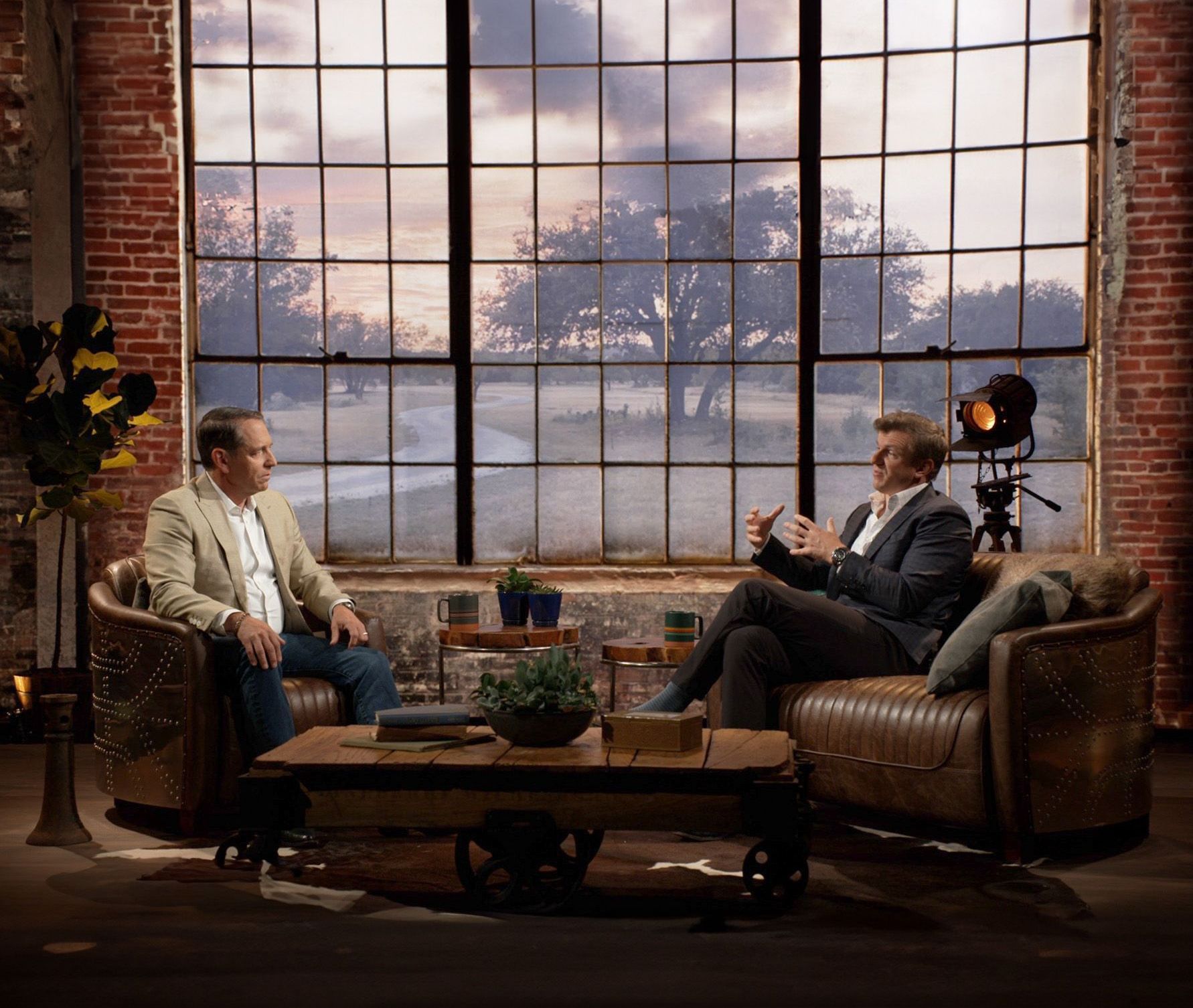 Two men are sitting in chairs and talking in front of a large window.