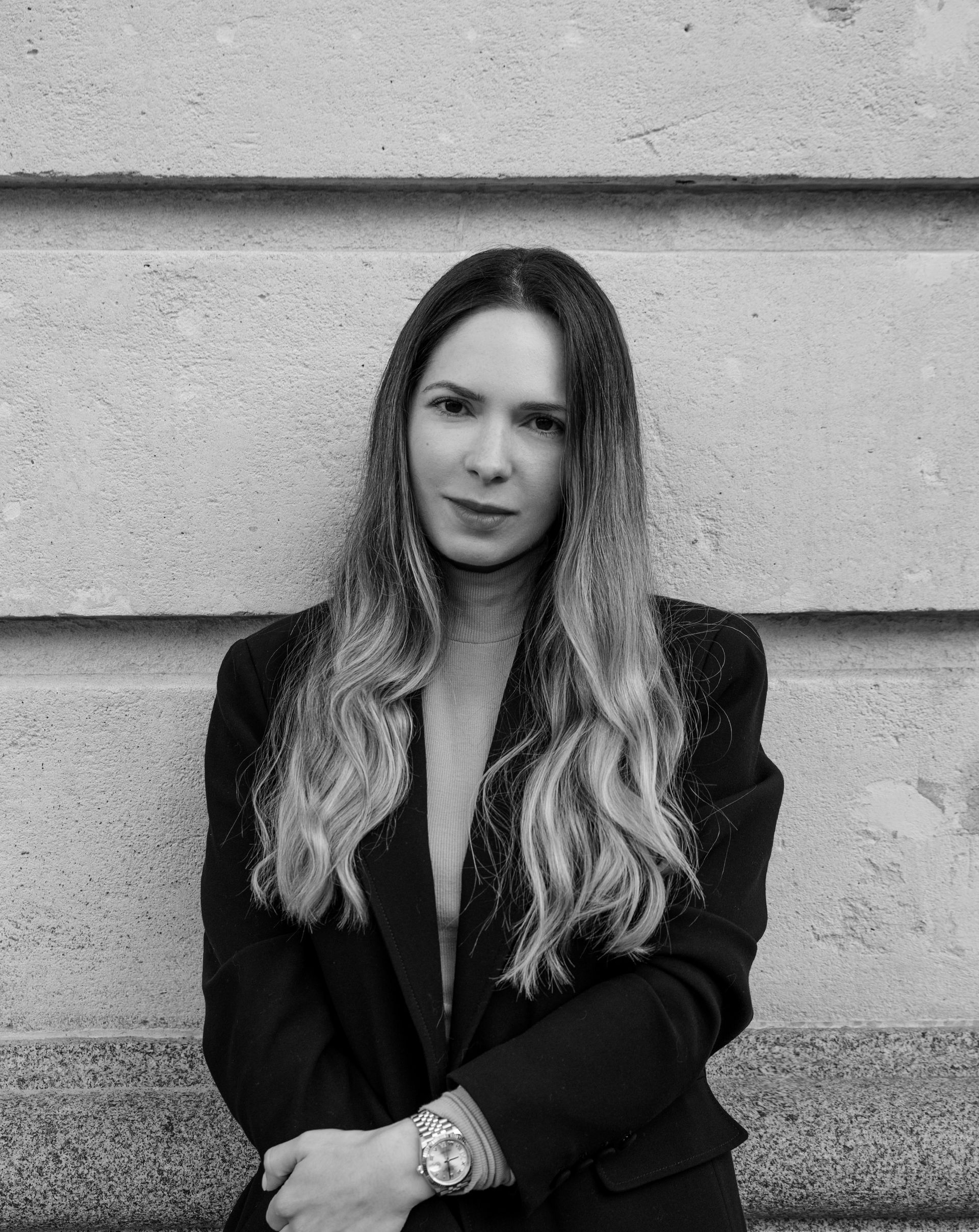 Woman with long hair, in blazer, leaning against textured wall.