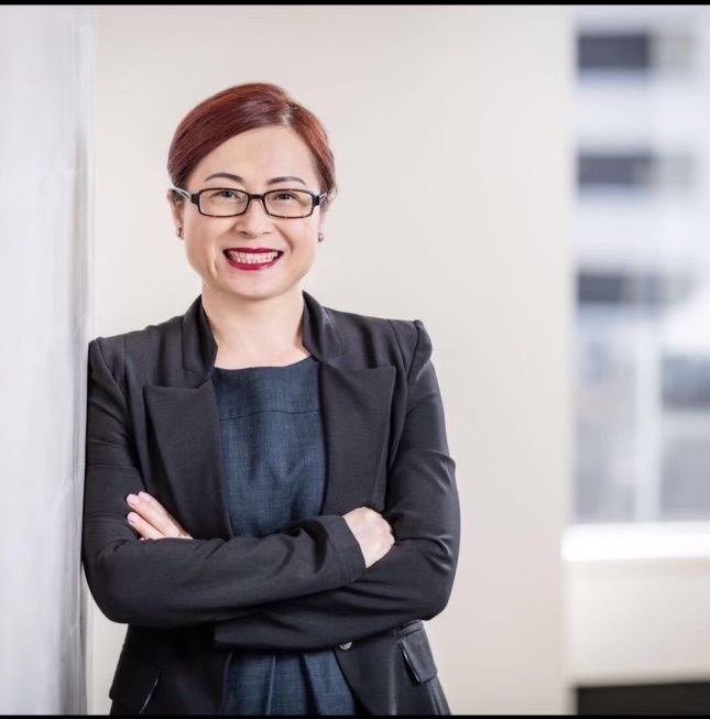 Woman with glasses, arms crossed, smiling, wearing a black blazer and dark top, leaning against a wall.