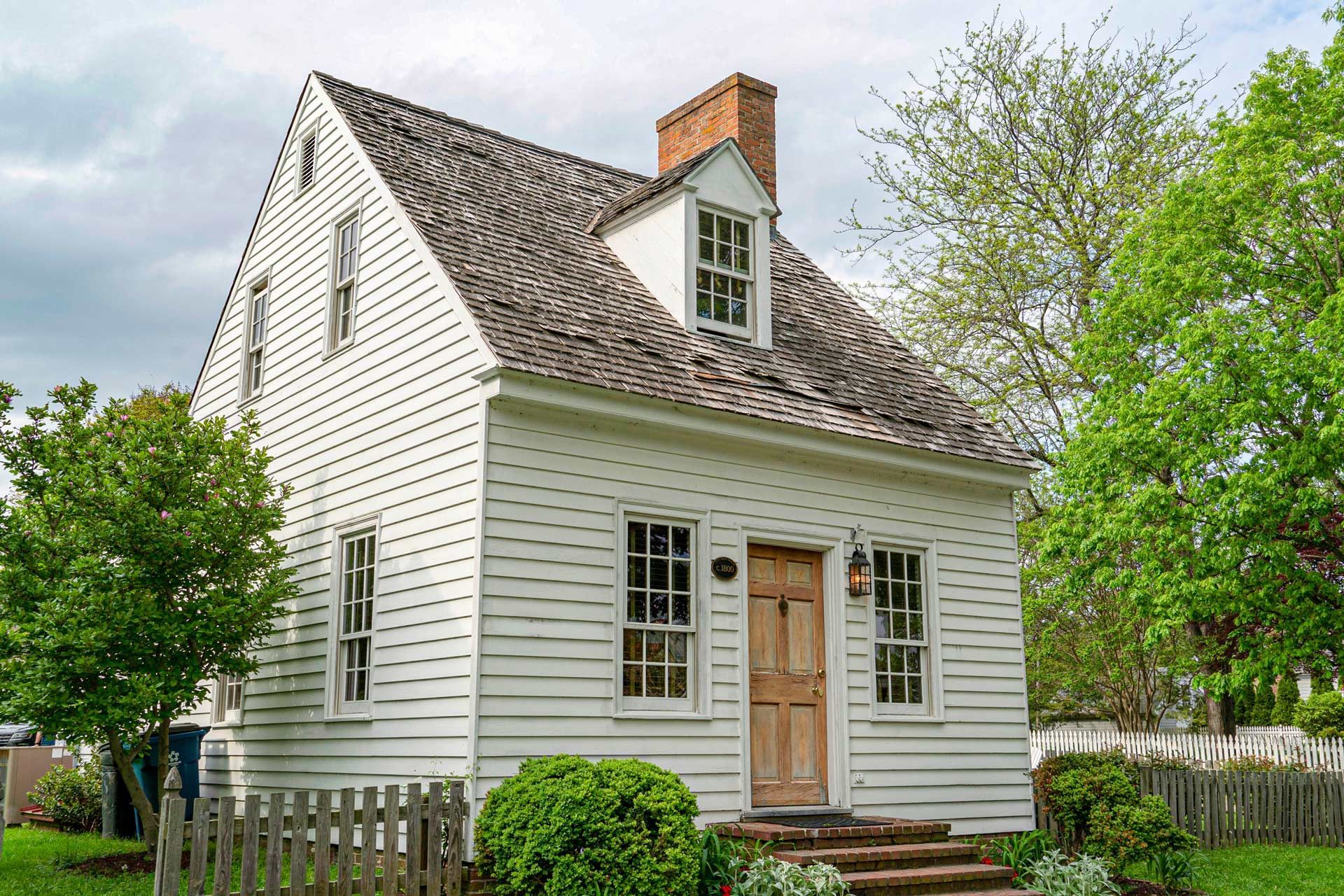 White, two-story house with a gabled roof and a brick chimney. Wooden siding, small windows, and a wooden front door.