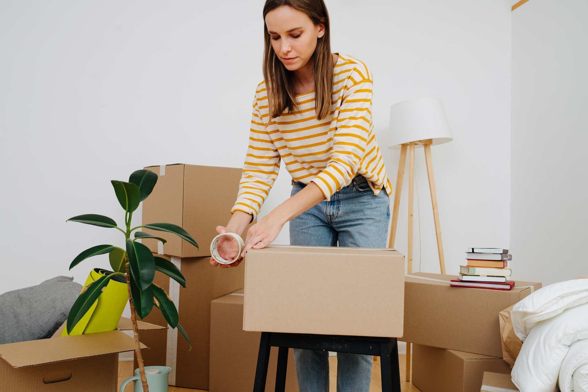 Woman sealing a cardboard box with tape in a room filled with moving boxes.