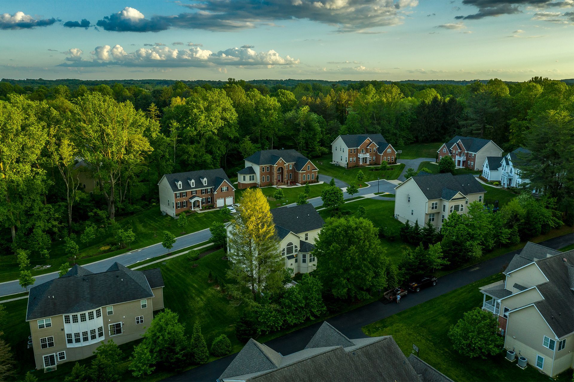 Aerial view of suburban homes surrounded by green trees and grass under a blue and cloudy sky.
