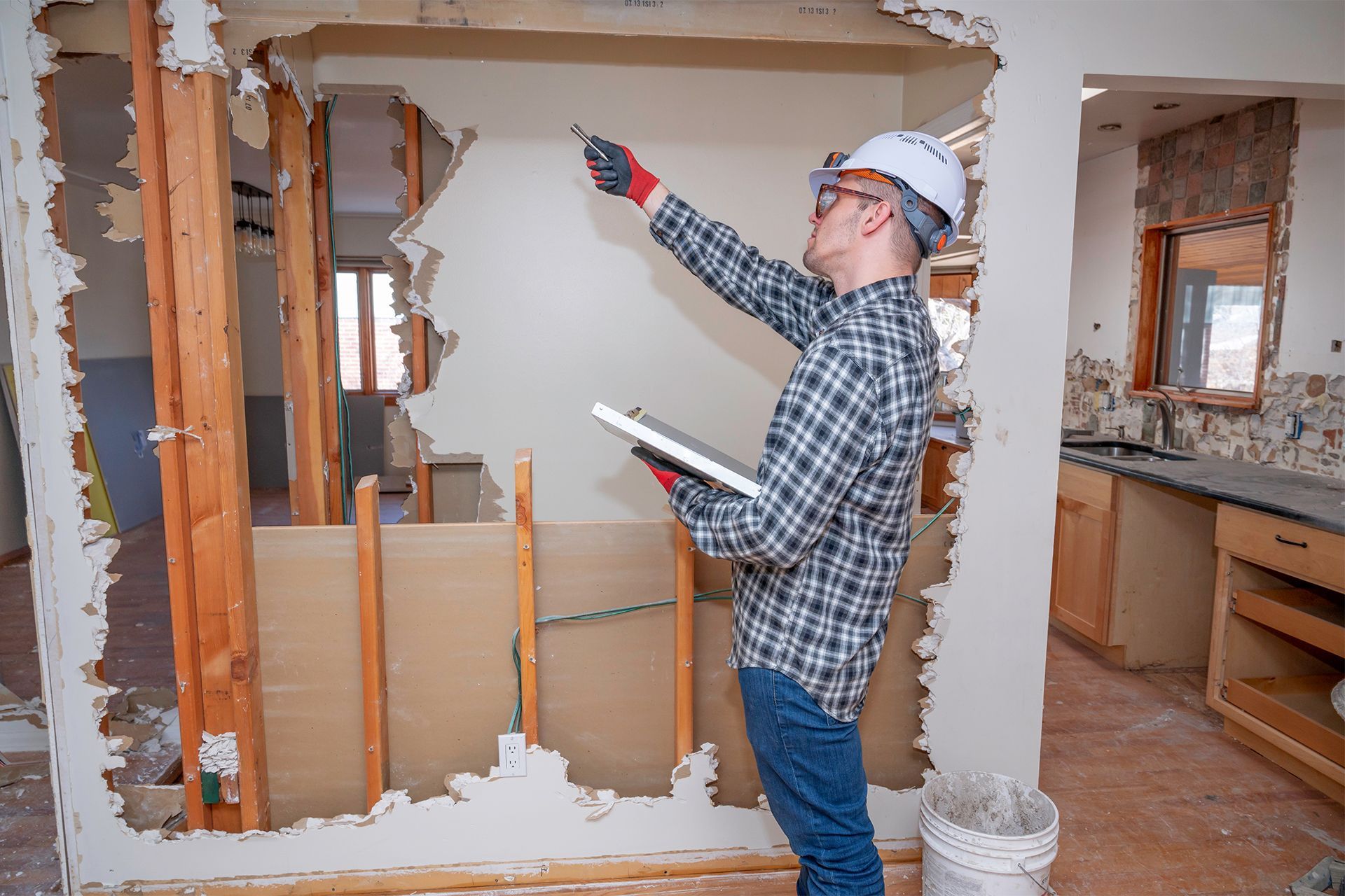 Construction worker in hard hat inspecting wall damage, holding clipboard and trowel. Interior shot.