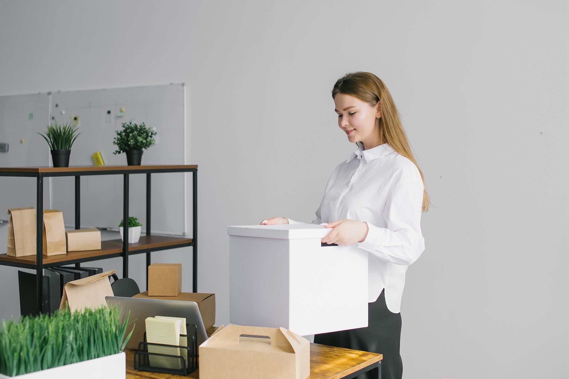 Woman holding a white box in an office, smiling. Boxes and plants on a shelf and desk.