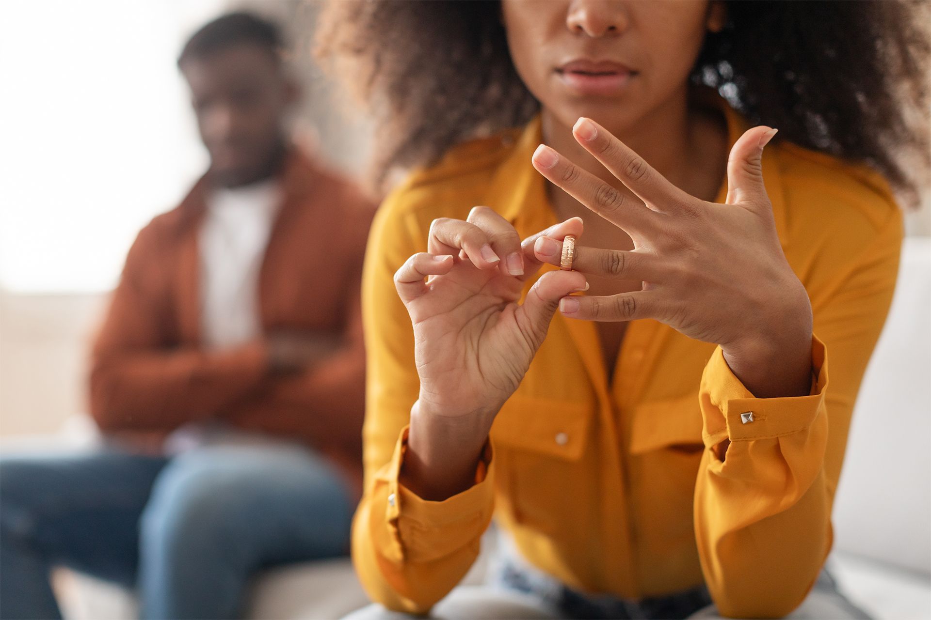 Woman removing ring, man in background; strained expressions, living room setting.