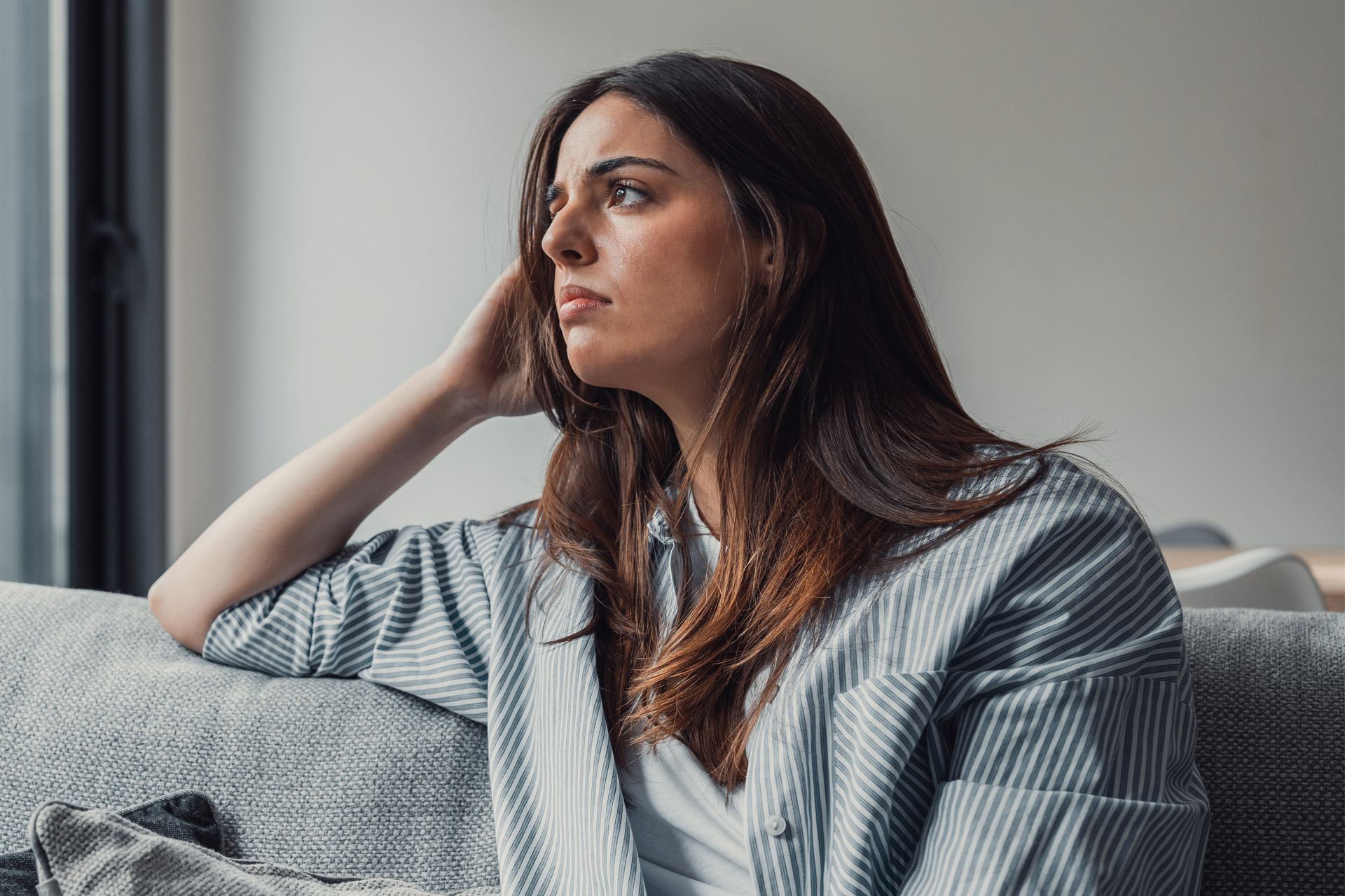 Woman sitting on a couch, gazing out a window, looking thoughtful.