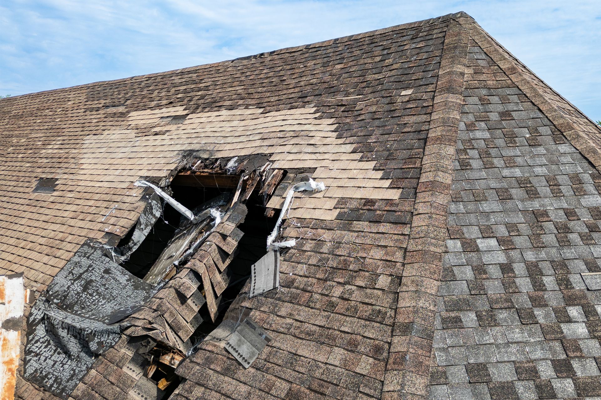 Damaged, weathered rooftop with a large hole revealing structural supports and exposed shingles.