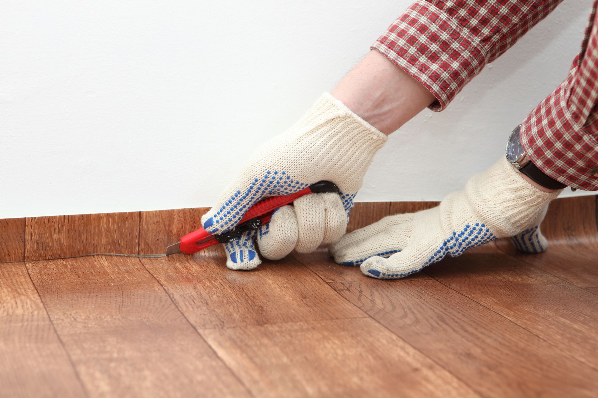 Person wearing gloves uses a utility knife to cut near wooden baseboard and flooring.
