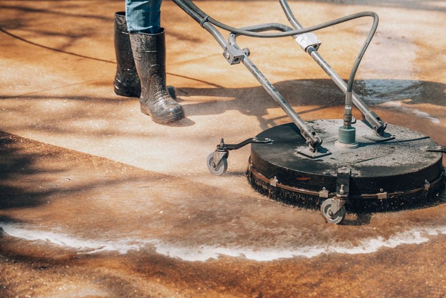 A person in rubber boots uses a circular pressure washer to clean a dirty concrete surface.