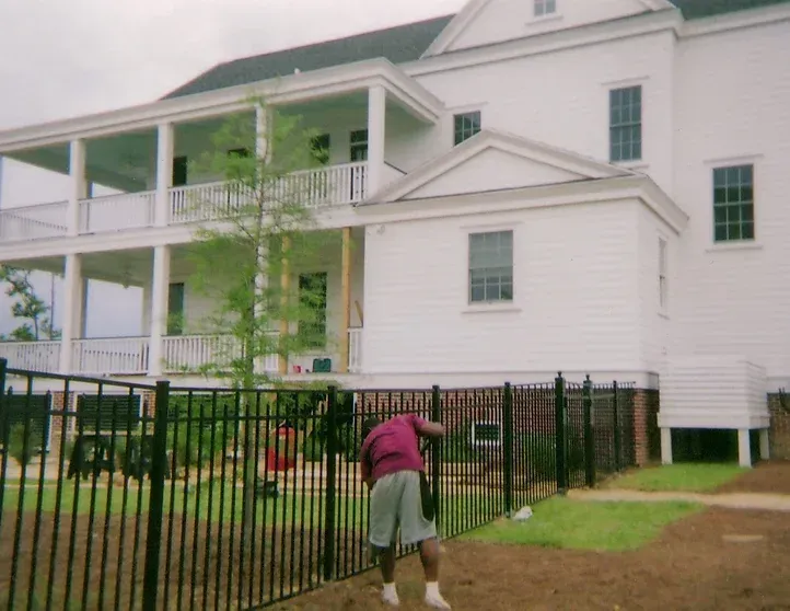 A man in a pink shirt is standing in front of a large white house