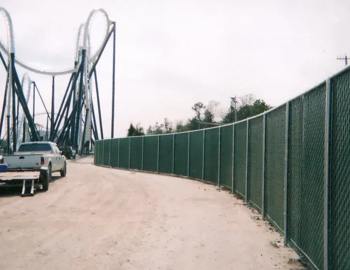 A chain link fence along a dirt road with a roller coaster in the background