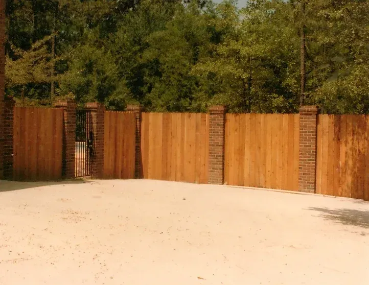 A wooden fence surrounds a sandy area with trees in the background.