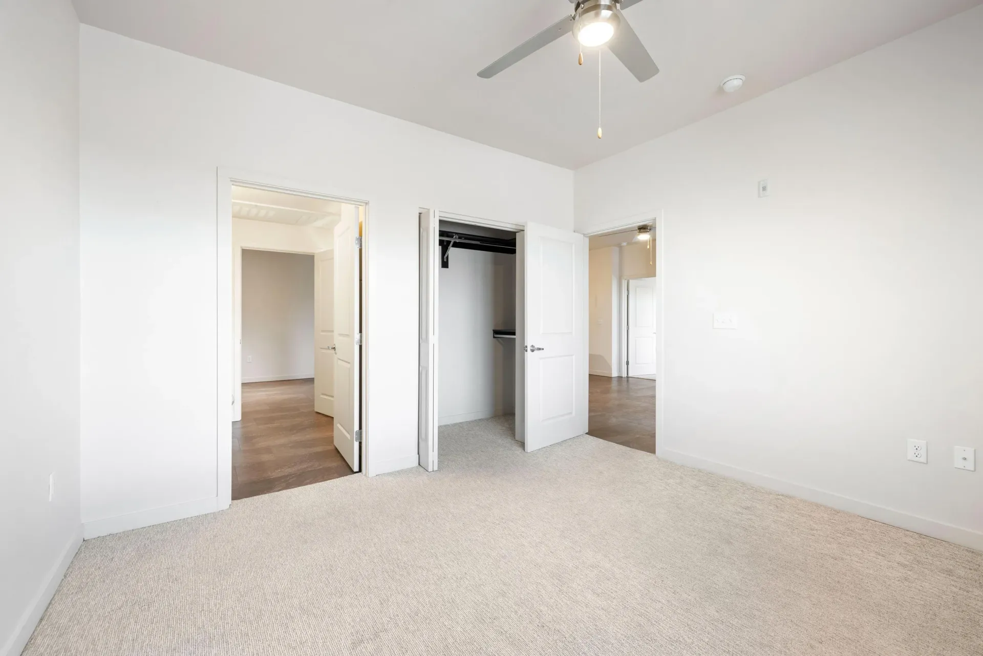 Bright bedroom with white walls, beige carpet, a ceiling fan, and open closet doors.