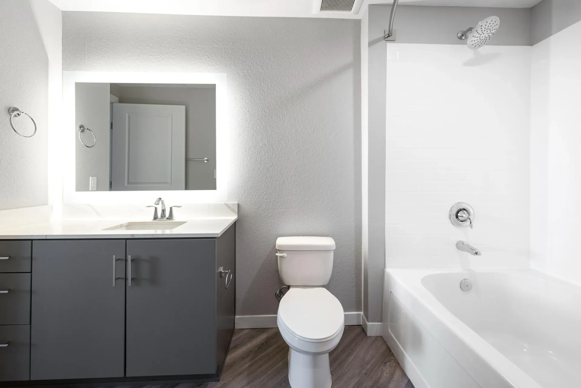 Modern apartment bathroom with grey vanity, backlit mirror, toilet, and white tub.