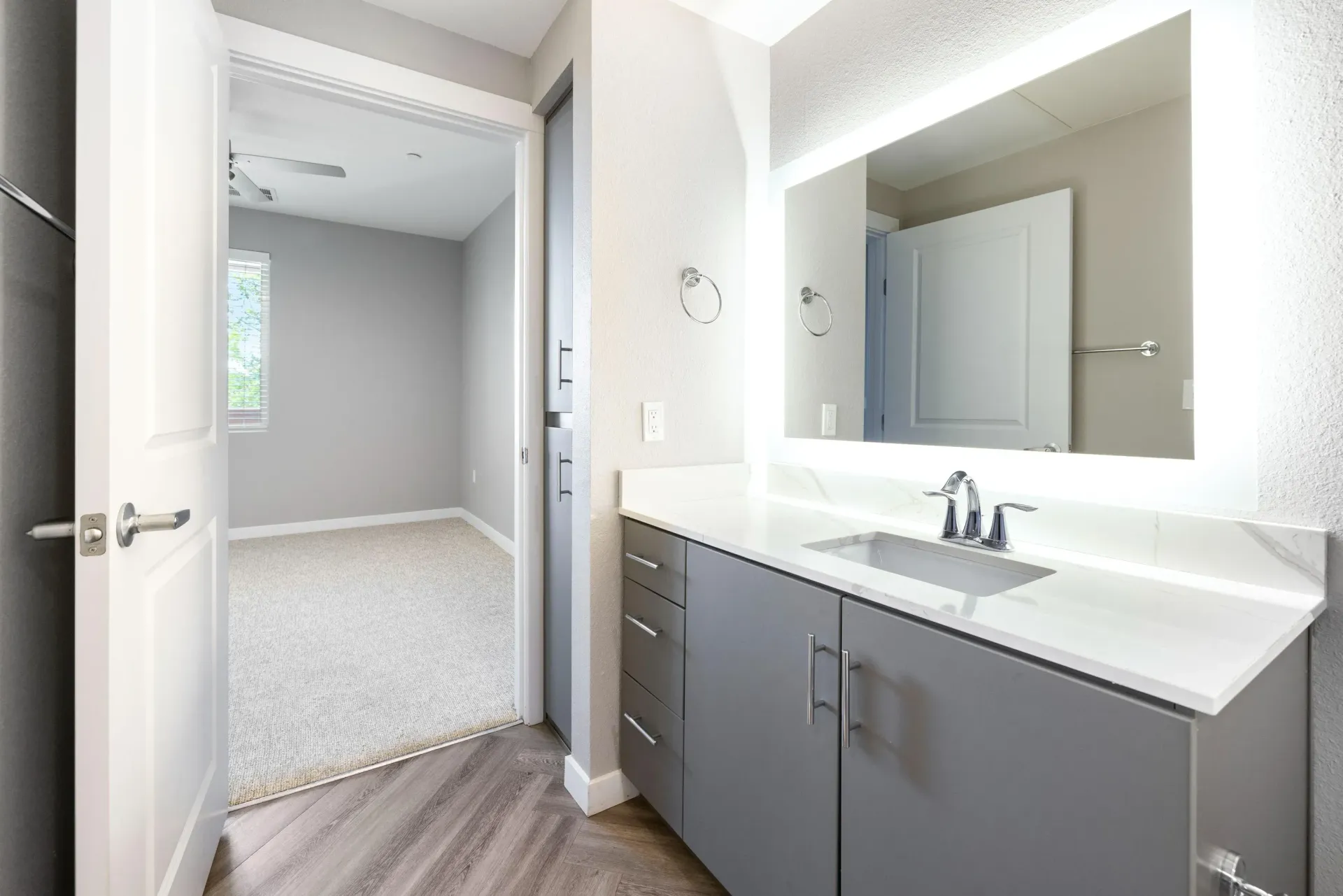 Bathroom vanity with gray cabinets, white countertop, and a backlit mirror.