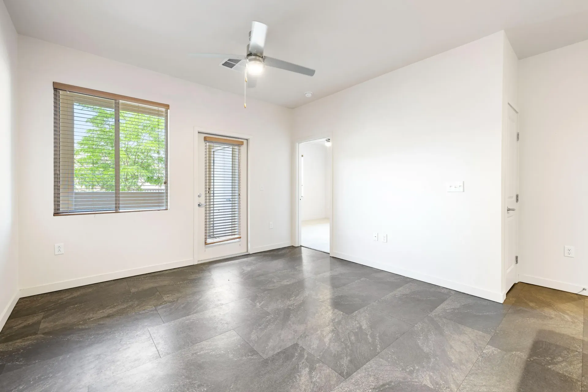 Empty living room with tile floor, white walls, ceiling fan, and door with blinds.
