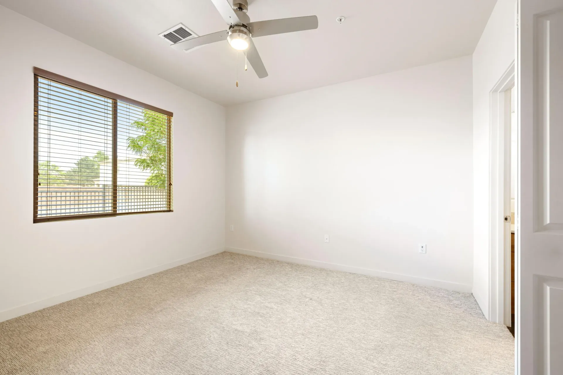 Empty bedroom with beige carpet, white walls, a window with wooden blinds, and a ceiling fan.