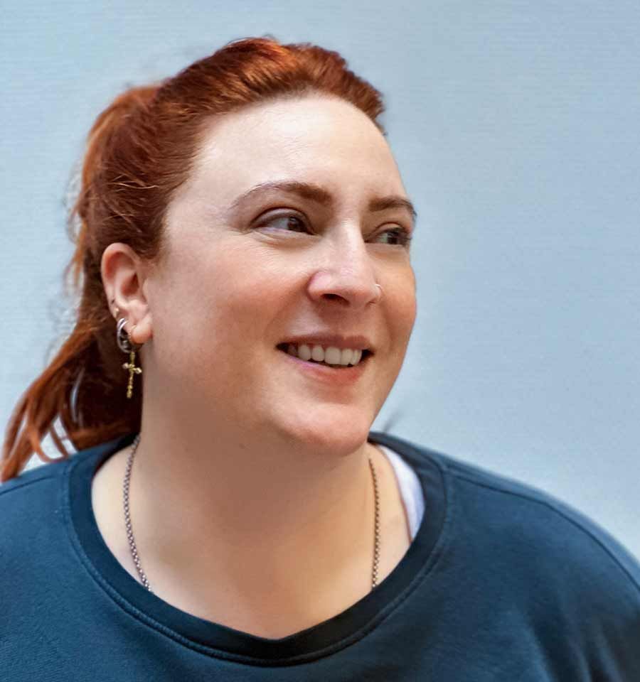 Une femme aux cheveux roux porte un pull bleu et des boucles d'oreilles et sourit.