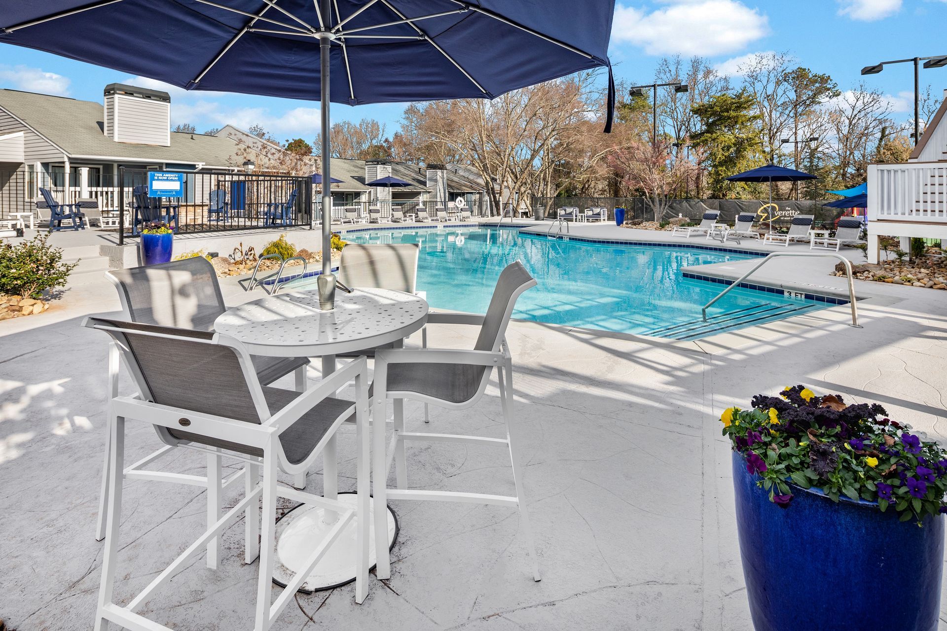 Poolside patio with blue umbrella, tables, and chairs. Pool in the background, sunny day.