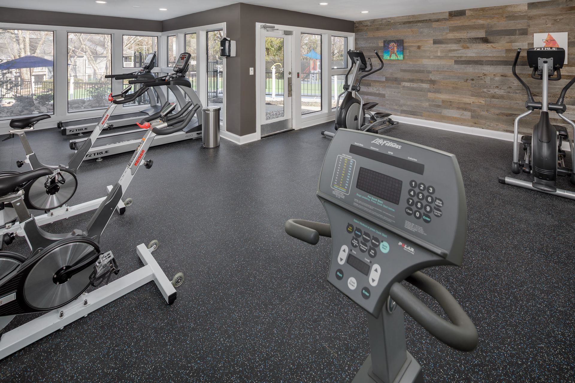 Gym with exercise equipment, including treadmills and stationary bikes, with wood-paneled wall and glass doors.