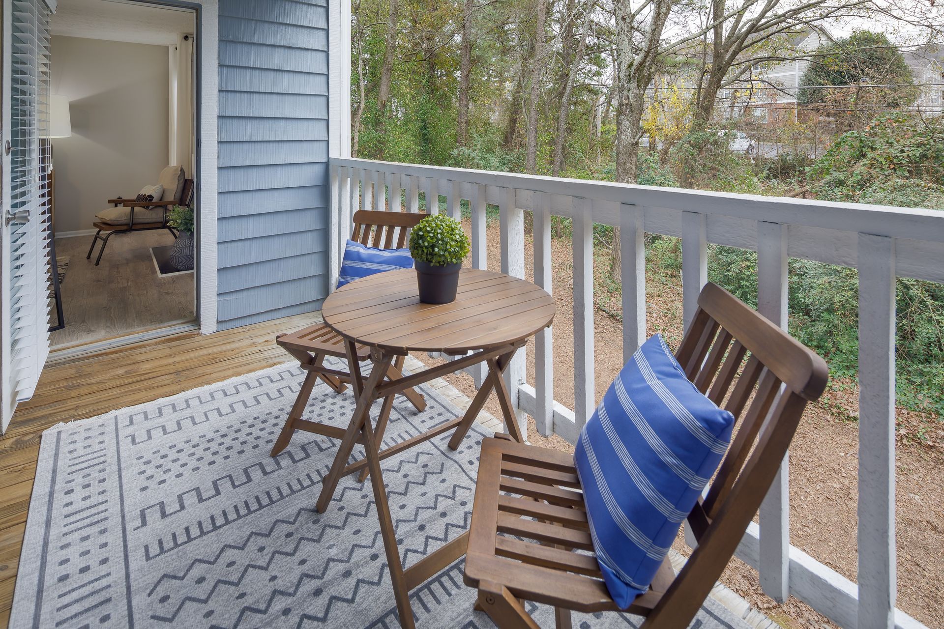 Wooden balcony with a small table, chairs, and blue-striped pillows, overlooking a yard.