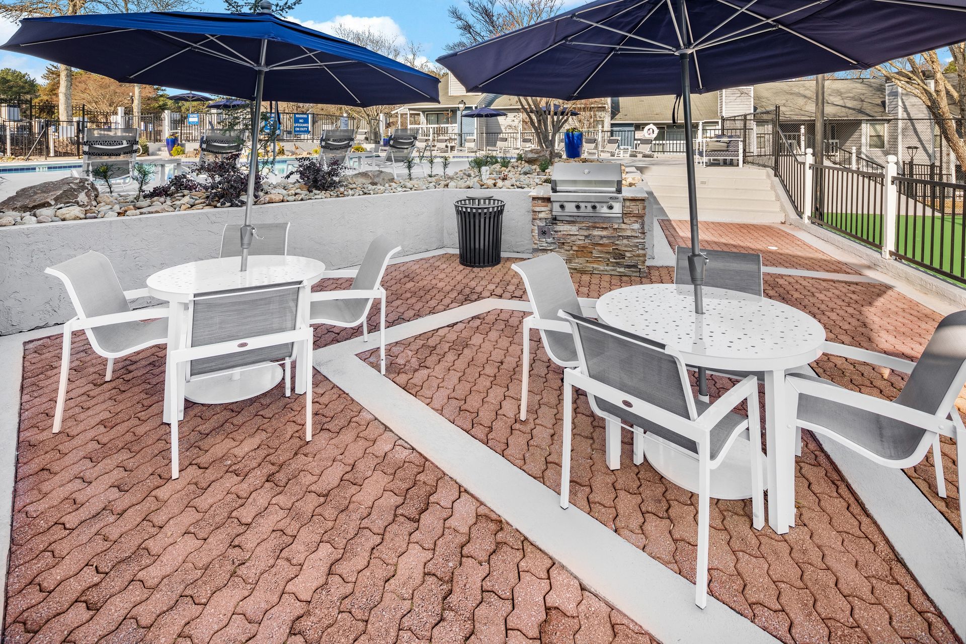 Outdoor patio with tables, chairs, umbrellas, and a grill. Brick flooring with white accents near a pool.