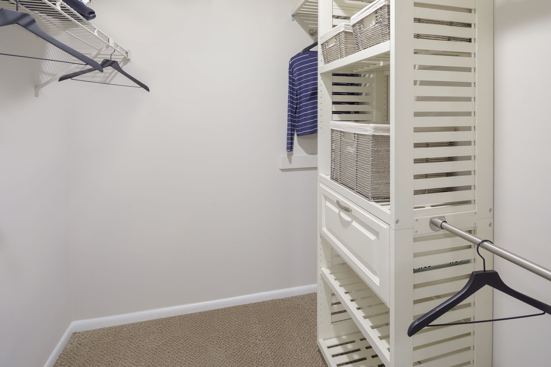 White closet with shelves, baskets, and hanging rod.  Clothes hang on a rack.  Neutral carpet.
