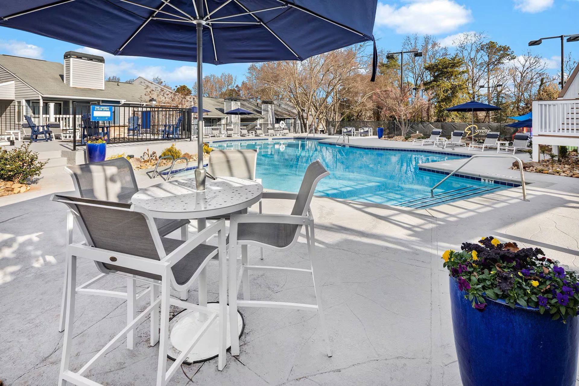 Poolside view with tables, chairs, and blue umbrella overlooking a pool on a sunny day.