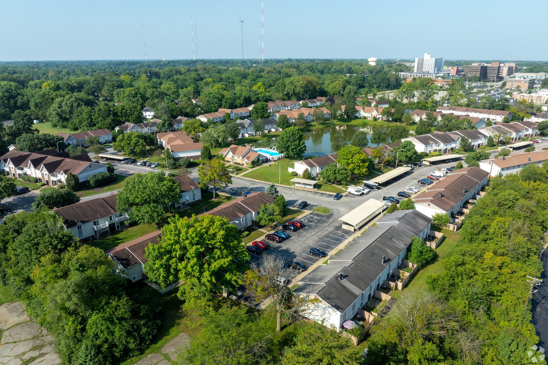 Aerial view of a residential neighborhood with townhomes, trees, a pond, and distant buildings under a blue sky.