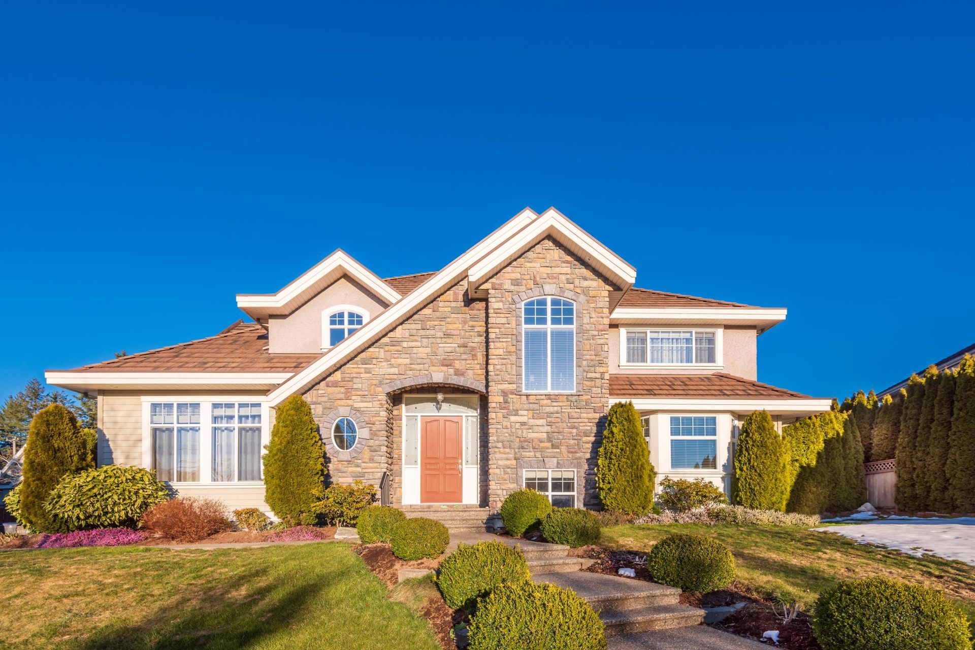 Stone-faced house with brown roof, orange door, and blue sky. Lush landscaping with green shrubs and a pathway.